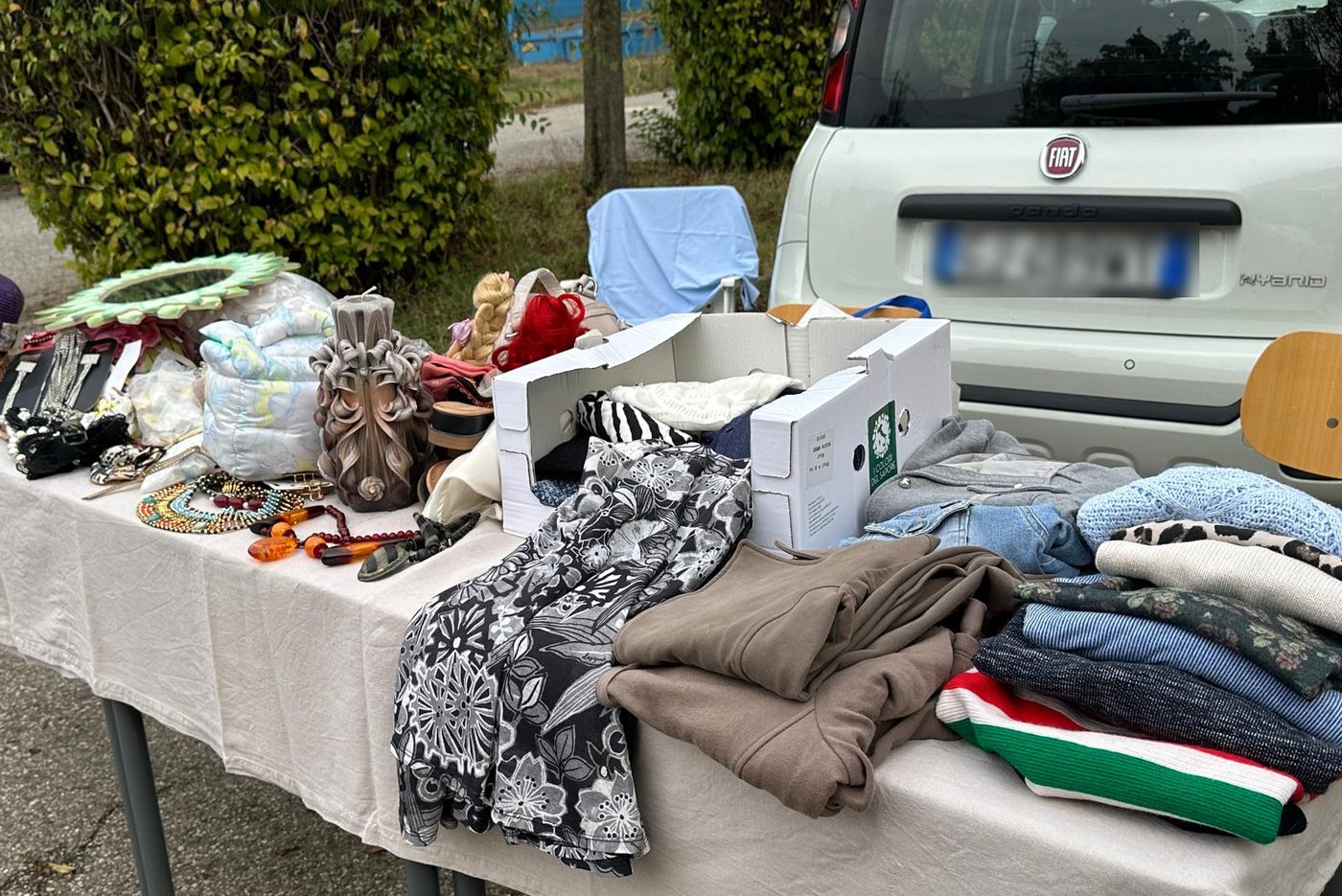 Un banchetto con collane, una candela scolpita e pile di vestiti piegati davanti a un’auto